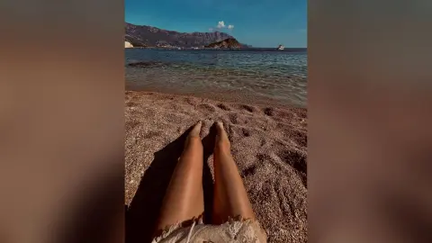 Person relaxing on a sunny beach with legs stretched out toward the blue ocean and distant mountains.