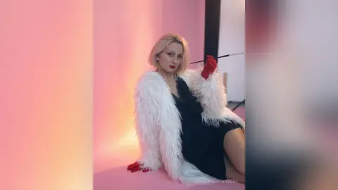 Glamorous blonde woman in a black dress and white faux fur coat posing against a pink background.