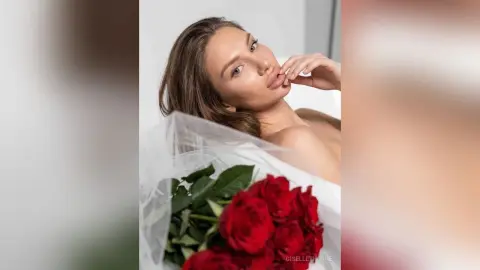 Beautiful woman with long brown hair relaxing in white sheets next to a bouquet of red roses in a bright room.