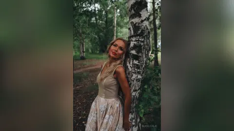 AliceLeslie — AliceLeslie leans against a birch tree in a sleeveless beige dress in a forest setting.