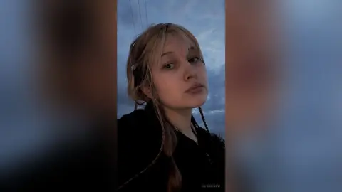 EloiseGow looks into the camera with blonde hair and braids against a cloudy blue sky.
