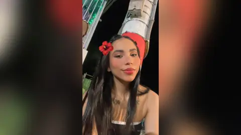 Smiling young woman with long dark hair and a red flower accessory in a nighttime outdoor setting.