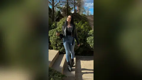 Young woman in black leather jacket and blue jeans holding coffee on outdoor stone stairs in a sunny park.