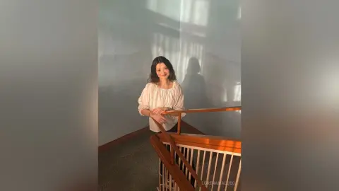 Smiling woman in a white blouse standing on a wooden staircase in a bright, minimalist room.