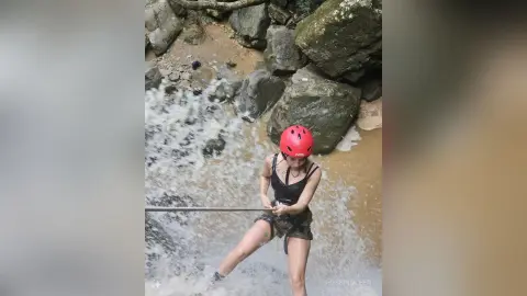 Adventurous woman in black activewear and red helmet rappelling down a rocky river waterfall.