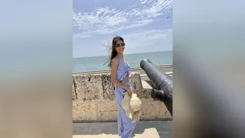 Smiling woman in a blue striped summer outfit holding a straw hat by the ocean on a sunny day.