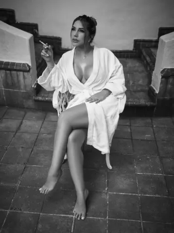 Elegant woman in a white bathrobe sitting on stone stairs with a cigarette in a moody black and white setting.