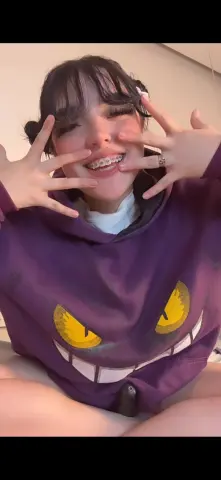 Smiling young woman with dark hair and braces wearing a purple graphic hoodie in a cozy indoor setting.