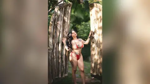 Curvy woman in a red bikini posing by rustic wooden gates in a lush tropical garden setting.