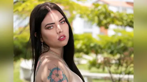 Beautiful woman with long dark hair and floral tattoos posing outdoors in a sunny garden setting.