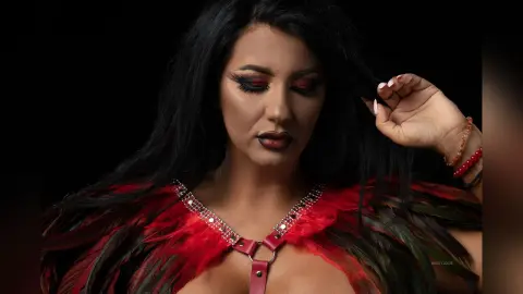 MissyJolie poses with her eyes downcast, wearing a red and black feathered collar with silver stud detailing.