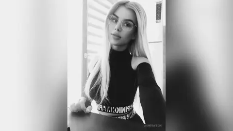 Blonde woman in black turtleneck top and glasses posing with a moody aesthetic in a bright indoor setting.