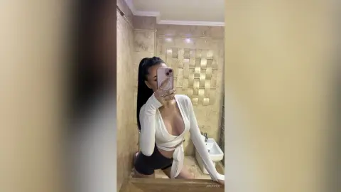 AriaVex poses for a mirror selfie wearing a white wrap top and black shorts in a marble bathroom.