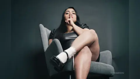 Elegant woman in a black dress and white socks sitting on a grey armchair against a dark studio background.