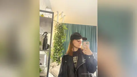 Young woman in a black leather jacket and baseball cap taking a mirror selfie in a cozy, modern room.