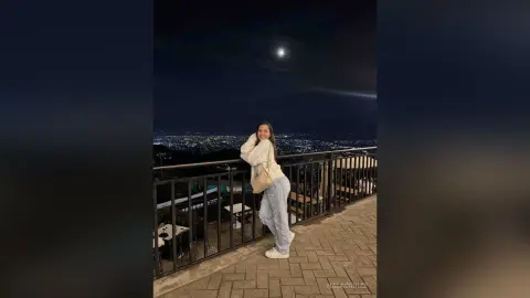 Smiling woman in a white sweater and jeans standing on a balcony overlooking a city skyline at night.