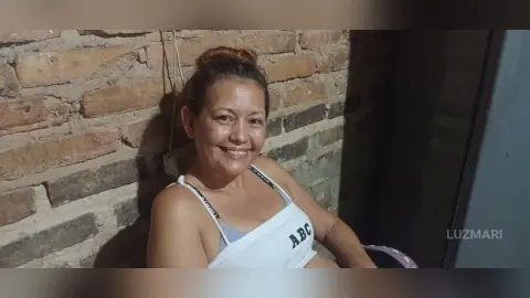 Smiling woman in a white tank top sitting against a rustic brick wall in a warm, casual setting.