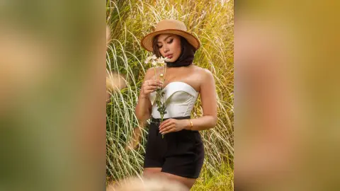 Stylish woman in a white strapless top and straw hat holding white flowers in a sunny meadow.