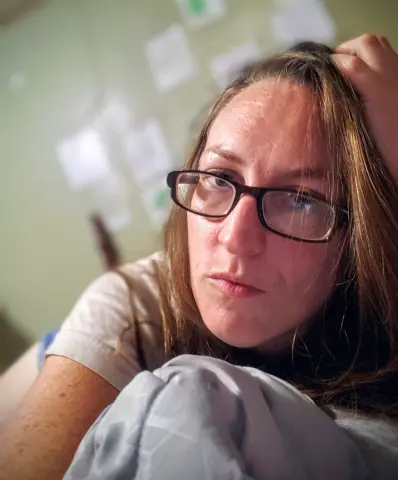 Thoughtful woman with glasses relaxing in a cozy indoor setting wearing a casual beige t-shirt.