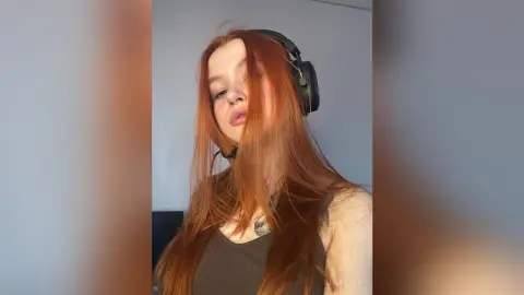 Redheaded woman wearing black headphones and a dark tank top in a minimalist indoor setting.