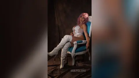 Elegant woman with pink hair in white lace bodysuit and tall boots sitting on a blue vintage chair.