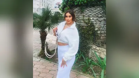 Stylish woman in a white fuzzy sweater and skirt holding sunglasses in a lush outdoor garden setting.