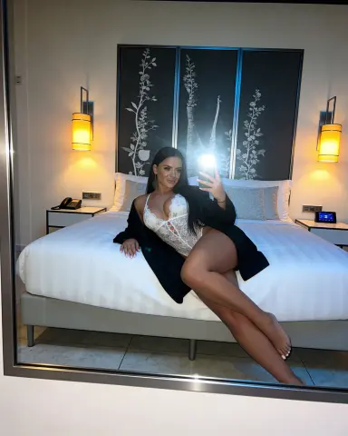 Woman in white lace lingerie and black robe sitting on a white bed in a modern bedroom.