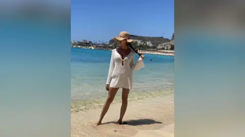 Woman in white knit mini dress and straw hat standing on a sunny tropical beach by turquoise ocean water.