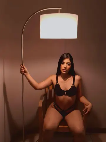 Young woman in black lingerie sitting by a floor lamp in a moody, dimly lit room.