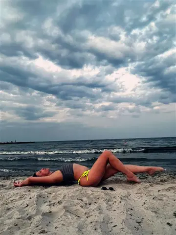 Woman in a black striped bikini relaxing on a sandy beach under a dramatic cloudy sky.