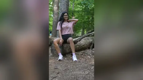Smiling young woman in a pink t-shirt and black shorts sitting on a log in a lush green forest.
