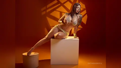 Glamorous woman in gold bikini and jewelry sitting on a white pedestal against a warm sunset background.