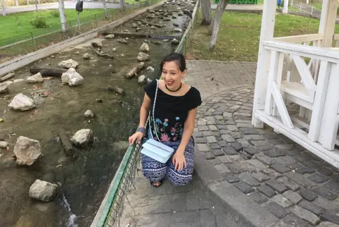 Smiling woman in a black floral top and patterned pants sitting by a rocky stream in a peaceful park.