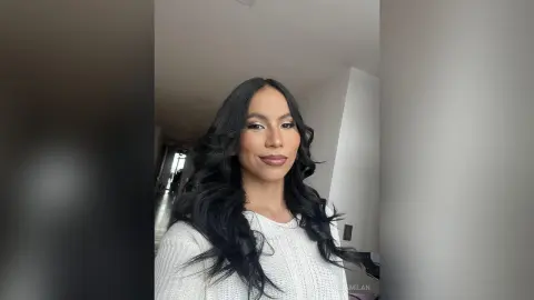 Smiling woman with long wavy black hair wearing a white knit sweater in a bright modern room.