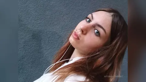 Young woman with nose piercings and long brown hair posing against a textured grey wall in a moody portrait.