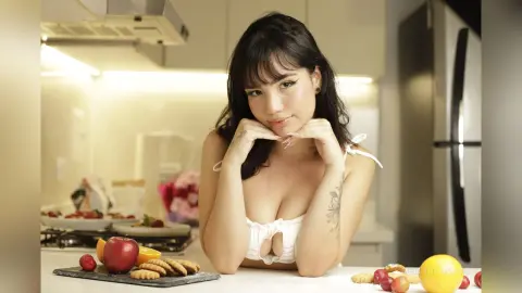 Young woman in a white bikini top smiling in a bright kitchen setting with fresh fruit on the counter.