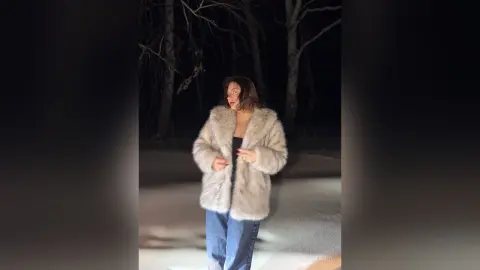 AnnyFraid stands in a snowy forest at night, wearing a thick, light-colored fur coat and blue jeans.