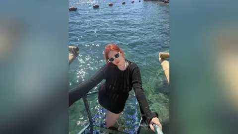 Red-haired woman in a black long-sleeve dress and white sunglasses relaxing by the sunny blue ocean.