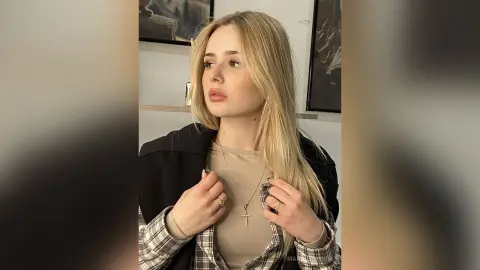 Blonde woman in a beige top and plaid shirt looking away in a bright indoor setting.