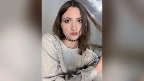 AriaLush wearing a grey long-sleeve shirt and a black choker, looking at the camera with her dark hair falling over her shoulder.