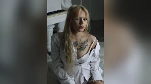 Blonde woman in a white button-down shirt sitting by a piano in a moody, cinematic indoor setting.