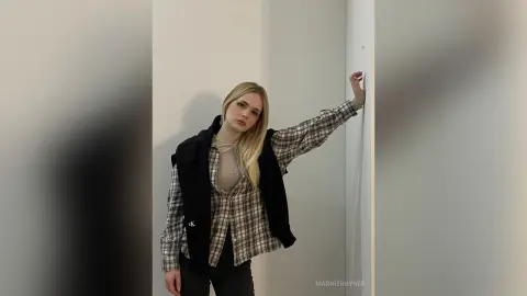 Blonde woman in a plaid shirt and black vest posing in a minimalist white studio setting.