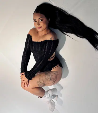 Young woman with long dark hair in a black knit crop top and shorts posing on a white studio background.