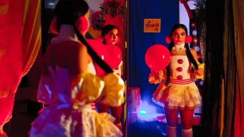 Young woman in a white and yellow clown costume with red pom-poms posing in a colorful circus-themed room.