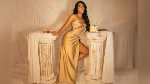 Elegant woman in a gold satin gown posing with a birthday cake between white Greek pillars in a dreamy studio.