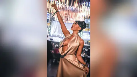 Dolce poses with one arm raised, wearing a gold satin halter dress with a deep plunging neckline.