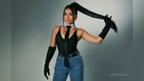 Stylish woman in a black bodysuit, tie, and long gloves posing against a grey studio background.