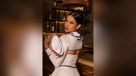 JuliahLewis poses in a white sailor-style top with blue trim, looking over her shoulder with her dark hair pulled back in a sleek bun.