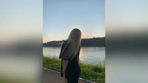 Blonde woman in a black dress looking at a calm lake during a peaceful sunset.