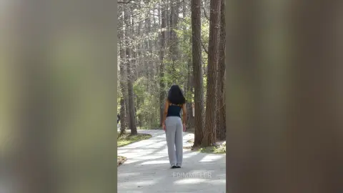 Woman in a black tank top and grey trousers walking through a serene, sunlit forest path.